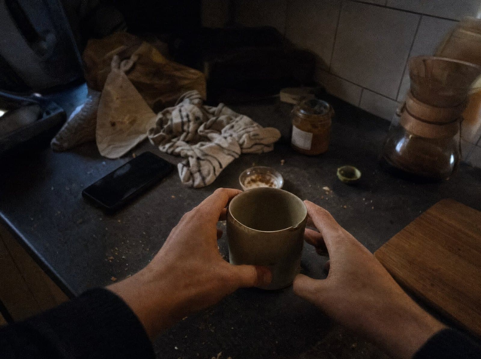 First-person view of two hands lifting a mug from a cluttered kitchen counter.